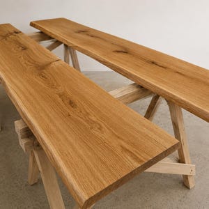 Könnte beinhalten: Zwei lange, rechteckige Holzbohlen mit natürlichen Baumkanten und sichtbarer Holzmaserung. Jede Bohle wird von einem hellen Holzbock getragen und weist eine warme, goldbraune Oberfläche sowie ausgeprägte Astlöcher im Holz auf.