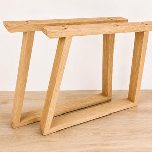 Peut inclure: Deux pieds de meuble en bois clair avec une structure trapézoïdale. Chaque pied présente une barre de montage supérieure horizontale avec des trous pré-percés, un support vertical incliné et une base horizontale plate en bois naturel.