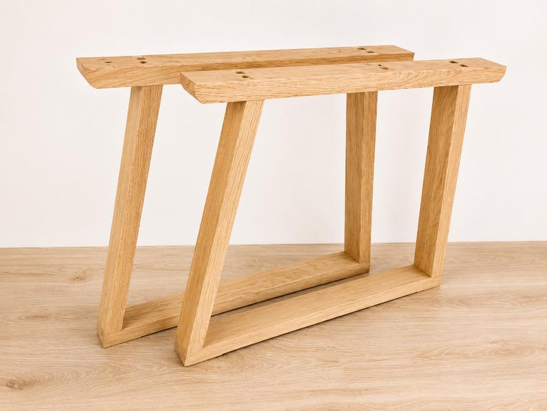 May include: Two light-colored wooden furniture legs with a trapezoidal frame design. Each leg features a horizontal top mounting bar with pre-drilled holes, a slanted vertical support, and a flat horizontal base piece made of natural wood grain.