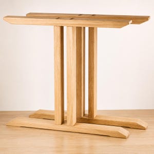 Op de afbeelding: Een lichtgekleurde houten tafelonderstel met twee parallelle horizontale steunbalken verbonden door vier verticale palen. De structuur is gemaakt van natuurlijk hout met een zichtbare nerf en toont een stevig frame met schuine uiteinden.