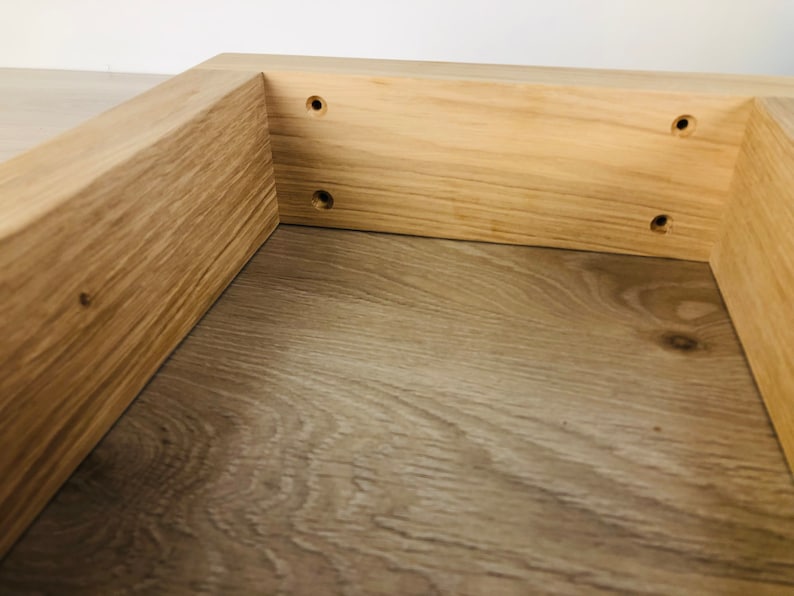 May include: A light-colored wooden frame with a natural grain pattern, featuring four pre-drilled circular holes along the inner side panel. The structure is constructed from solid wood planks joined at a right angle, showcasing a smooth finish.