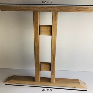 Puede incluir: Una base de mesa de madera de color marr&oacute;n claro con una parte superior rectangular y una base ancha y plana, ambas de 580 mm de ancho. La estructura cuenta con dos soportes verticales conectados por dos peque&ntilde;os bloques cuadrados, con veta de madera natural.
