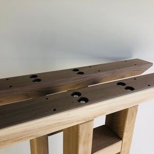 May include: Two parallel wooden furniture legs featuring a dark brown walnut top section and a lighter oak base. The top surfaces have pre-drilled circular holes and recessed metal hardware for mounting, showcasing a dual-tone wood construction.