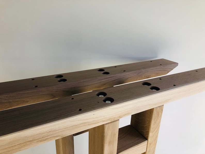 May include: Two parallel wooden furniture legs featuring a dark brown walnut top section and a lighter oak base. The top surfaces have pre-drilled circular holes and recessed metal hardware for mounting, showcasing a dual-tone wood construction.