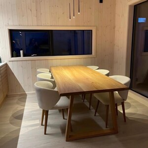 May include: A rectangular wooden dining table with a natural grain finish and a sturdy frame, accompanied by six upholstered chairs with light-colored fabric and wooden legs, positioned in a room with light-toned wood paneled walls.