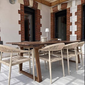 May include: A long rectangular wooden dining table with a live edge top and a sturdy trestle base, accompanied by several chairs featuring light beige woven rope seats and backrests, arranged on a light-colored patio under a wooden pergola.