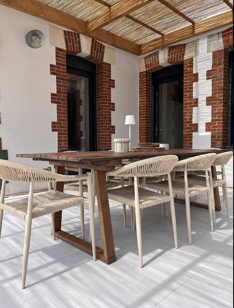 May include: A long rectangular wooden dining table with a live edge top and a sturdy trestle base, accompanied by several chairs featuring light beige woven rope seats and backrests, arranged on a light-colored patio under a wooden pergola.