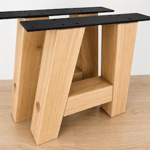Può includere: Due gambe per tavolo a forma di A realizzate in legno naturale di tonalità chiara con venature visibili. Ogni gamba presenta una trave di supporto orizzontale e una piastra di montaggio in metallo nero piatto sulla parte superiore.