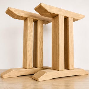Op de afbeelding: Twee lichtgekleurde houten tafelpoten met een dubbel kolomontwerp, verbonden door een horizontale bovenste steun en een brede basis. De natuurlijke houtnerf is zichtbaar door de hele structuur, met een lichtbruine afwerking en schuine randen.