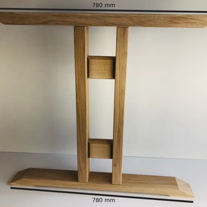 Puede incluir: Una mesa consola de madera de color marr&oacute;n claro con un tablero rectangular y una base ancha, ambos de 780 mm de ancho. La mesa tiene dos patas de soporte verticales conectadas por dos bloques de madera horizontales, creando una estructura estable.