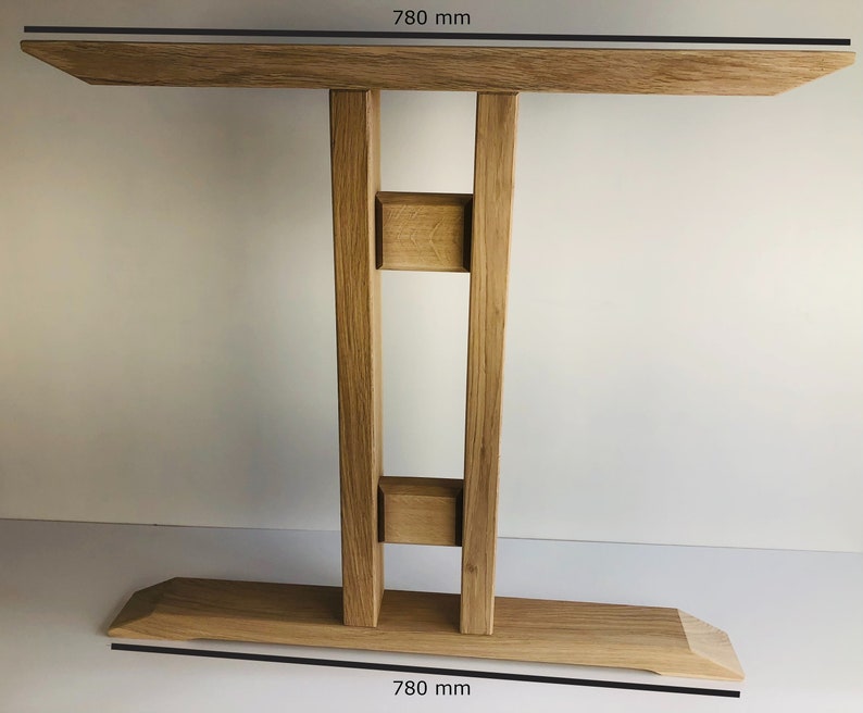 Puede incluir: Una mesa consola de madera de color marr&oacute;n claro con un tablero rectangular y una base ancha, ambos de 780 mm de ancho. La mesa tiene dos patas de soporte verticales conectadas por dos bloques de madera horizontales, creando una estructura estable.