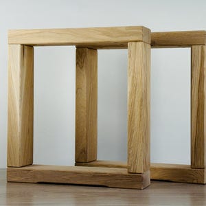 May include: Two rectangular table legs constructed from light-colored natural wood with a visible grain pattern. Each leg features a sturdy base, two vertical supports, and a horizontal top beam, forming a clean, open, and geometric frame structure.