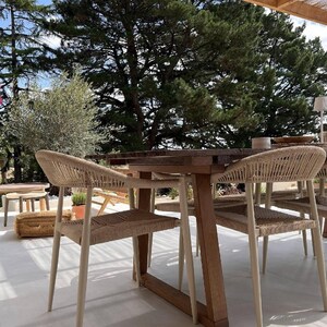 May include: A wooden dining table with a dark brown finish and four chairs featuring light beige woven rope seats and backrests. The chairs have tapered legs and a curved frame design, positioned around the table on a light-colored patio floor.
