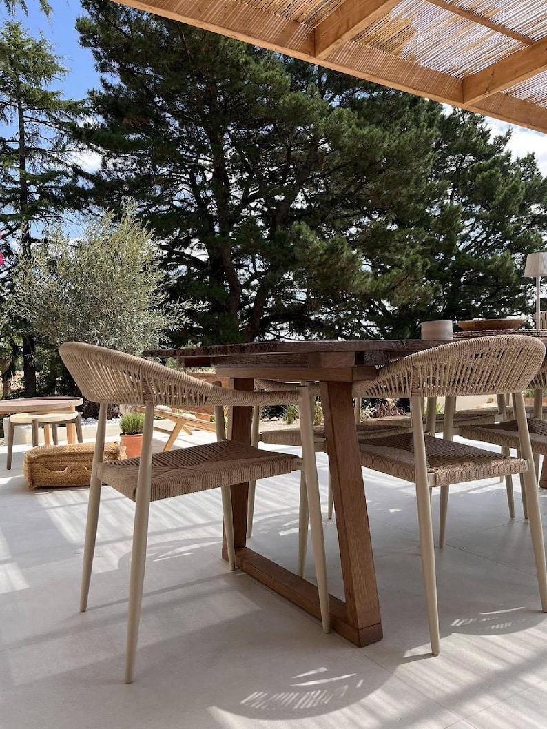 May include: A wooden dining table with a dark brown finish and four chairs featuring light beige woven rope seats and backrests. The chairs have tapered legs and a curved frame design, positioned around the table on a light-colored patio floor.