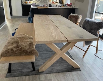 Op de afbeelding: Een grote houten eettafel met een natuurlijke afwerking en een kruisvormig onderstel. De tafel is voorzien van een bank en twee stoelen, allemaal met kunstvacht kussens.