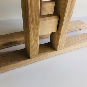 Puede incluir: Una base de mesa de madera de roble marr&oacute;n claro con dos postes de soporte verticales conectados por una barra transversal horizontal, apoyada sobre dos pies largos, planos y biselados. La veta natural de la madera es visible en la estructura.