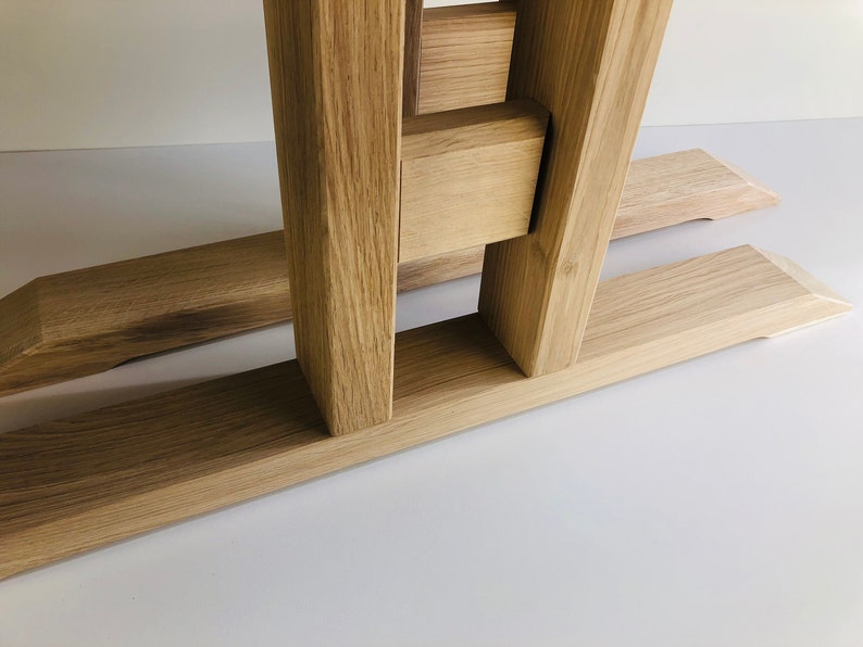 Puede incluir: Una base de mesa de madera de roble marr&oacute;n claro con dos postes de soporte verticales conectados por una barra transversal horizontal, apoyada sobre dos pies largos, planos y biselados. La veta natural de la madera es visible en la estructura.