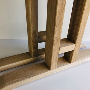May include: A light brown wooden table base featuring two vertical support beams connected by a horizontal crossbar, resting on two long, flat, beveled feet. The natural wood grain is visible throughout the structure, showcasing a light oak finish.