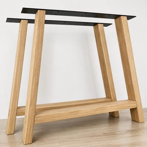 May include: A pair of table legs constructed from light-toned oak wood, featuring a trapezoidal frame design with a horizontal support beam near the base. Each leg assembly is topped with a flat, black metal mounting plate with pre-drilled holes.