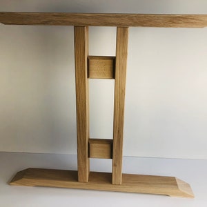 Puede incluir: Una mesa consola de madera de roble claro con una superficie rectangular, una base ancha y dos patas verticales de soporte conectadas por dos peque&ntilde;os bloques cuadrados. La veta de la madera es visible, mostrando un acabado natural claro.