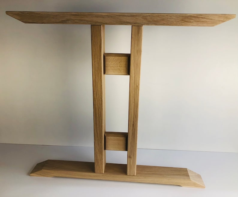 Puede incluir: Una mesa consola de madera de roble claro con una superficie rectangular, una base ancha y dos patas verticales de soporte conectadas por dos peque&ntilde;os bloques cuadrados. La veta de la madera es visible, mostrando un acabado natural claro.