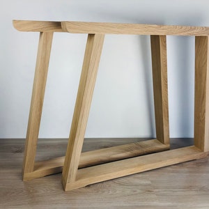 May include: Two light-colored wooden table legs featuring a trapezoidal frame design. Each leg consists of a horizontal base piece connected to a vertical support and an angled support, creating a sturdy structure with a natural wood grain finish.