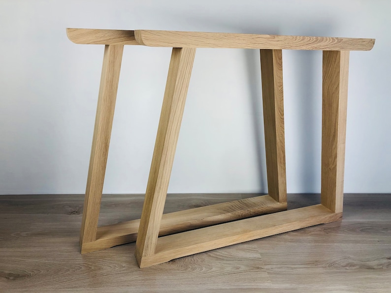May include: Two light-colored wooden table legs featuring a trapezoidal frame design. Each leg consists of a horizontal base piece connected to a vertical support and an angled support, creating a sturdy structure with a natural wood grain finish.