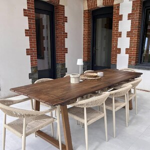 May include: A long rectangular wooden dining table with a dark brown finish and a trestle base, surrounded by six chairs featuring light beige woven rope seats and backrests, with a small white lamp and a woven placemat on the tabletop.