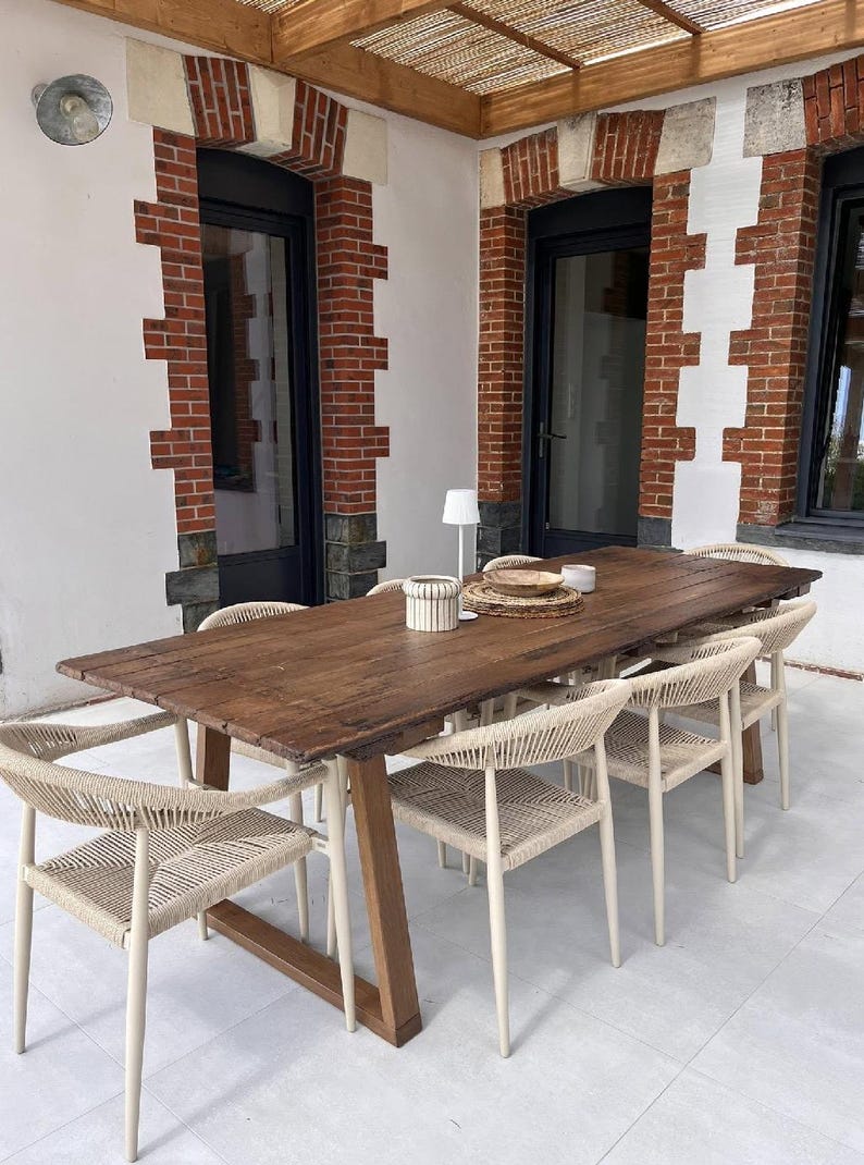May include: A long rectangular wooden dining table with a dark brown finish and a trestle base, surrounded by six chairs featuring light beige woven rope seats and backrests, with a small white lamp and a woven placemat on the tabletop.
