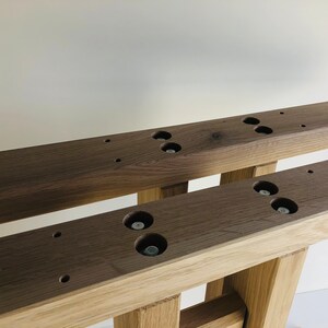 Puede incluir: Dos vigas de madera de color marr&oacute;n oscuro paralelas que presentan una serie de orificios circulares empotrados con cabezas de pernos met&aacute;licos visibles en su interior. Las vigas se apoyan sobre patas de madera verticales m&aacute;s claras.