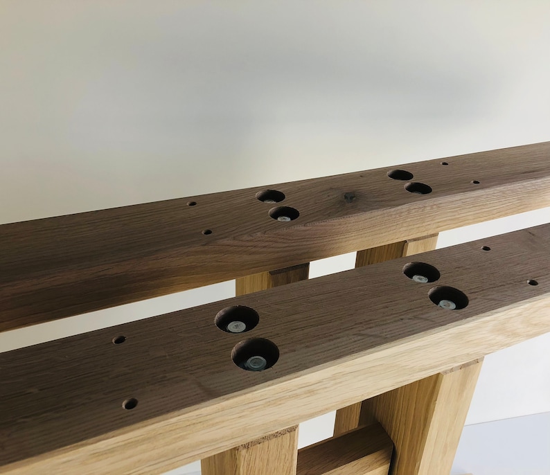 Puede incluir: Dos vigas de madera de color marr&oacute;n oscuro paralelas que presentan una serie de orificios circulares empotrados con cabezas de pernos met&aacute;licos visibles en su interior. Las vigas se apoyan sobre patas de madera verticales m&aacute;s claras.