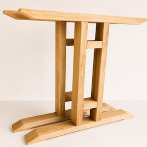 Peut inclure: Une base de table en bois brun clair composée d'une longue poutre de support horizontale supérieure, de deux pieds rectangulaires verticaux reliés par deux traverses horizontales et de deux pieds larges et inclinés à la base.
