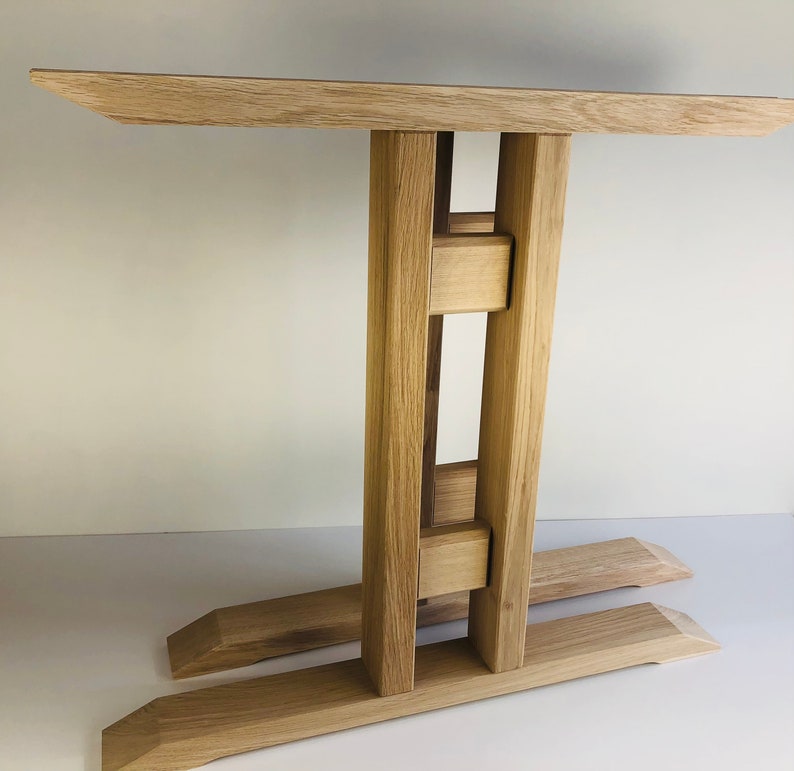 Puede incluir: Una mesa auxiliar de madera de color marr&oacute;n claro con una superficie rectangular sostenida por dos postes verticales conectados por dos travesa&ntilde;os horizontales, sobre una base compuesta por dos vigas paralelas con extremos biselados.