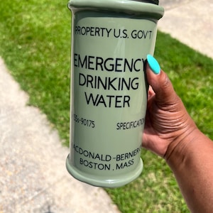 May include: A green reusable tumbler with a black lid and a straw. The tumbler has the text "PROPERTY U.S. GOV'T EMERGENCY DRINKING WATER 55-90175 SPECIFICATION MCDONALD-BERNIER BOSTON, MASS" printed on it.