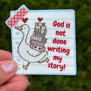 May include: A sticker featuring a white goose carrying a stack of books, with the text "God is not done writing my story!" in red. The sticker is on a blue background with a pink heart-patterned tape.