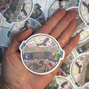 May include: A round sticker with a pink background and a green banner that says "protect planet earth". The sticker features a variety of animals and plants, including a bear, a whale, a turtle, a bird, and trees. The sticker is designed to promote environmental awareness.
