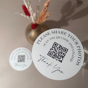 May include: Two white, circular stickers with black text and a QR code. The text reads "PLEASE SHARE YOUR PHOTOS, SCAN THE QR CODE AND UPLOAD, Thank You." A gold vase with dried flowers is in the background.
