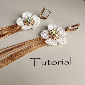 Puede incluir: Un par de pendientes hechos a mano con diseños de flores de cuentas blancas, una piedra preciosa central y borlas de cadena dorada. Los pendientes tienen ganchos para las orejas dorados. La palabra "Tutorial" está escrita en escritura negra.
