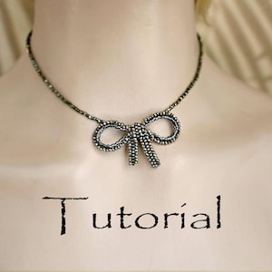 Könnte beinhalten: Eine Halskette mit einer Perlen-Schleife in filigranem Design. Die Halskette hat einen Anhänger in Form einer Schleife mit schwarzen und goldenen Perlen, und das Wort "Tutorial" ist darunter in schwarzer Schrift geschrieben.