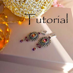 May include: A pair of silver earrings with a teardrop shape. The earrings are made with small beads and feature a large, iridescent red gemstone in the center. The earrings are on a pink surface with the word "Tutorial" written in black.