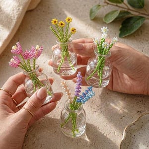 Puede incluir: Pequeños jarrones de cristal llenos de flores de cuentas en rosa, amarillo, blanco, lavanda, melocotón y azul. Las flores tienen tallos verdes y están dispuestas en jarrones de cristal transparente. Los jarrones son sostenidos por manos.