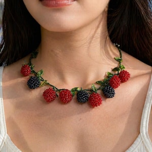 May include: A beaded necklace featuring red and black berry clusters with green leaves. The necklace is worn around the neck and is made of small, colorful beads. The berries are a prominent design element.