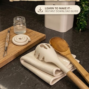 Könnte beinhalten: Digitales Tutorial für eine handwerkliche gefaltete Handtuch-Löffelablage. Das Bild zeigt eine weiße Keramik-Löffelablage, einen Holzlöffel und einen weißen Nudellöffel. Ein Holzschneidebrett mit Tonwerkzeugen und Tonspulen ist ebenfalls zu sehen.