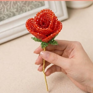 May include: A handcrafted rose sculpture constructed from small, vibrant red beads forming the petals and green beads creating the sepals. The stem is wrapped in a thin, metallic gold-colored wire, providing a slender and flexible support structure.