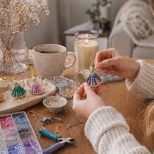 May include: A person is holding a beaded angel ornament with blue, pink, and gold accents. Other angel ornaments in various colors are displayed on a tray. Crafting supplies, including beads and tools, are visible on a wooden table.