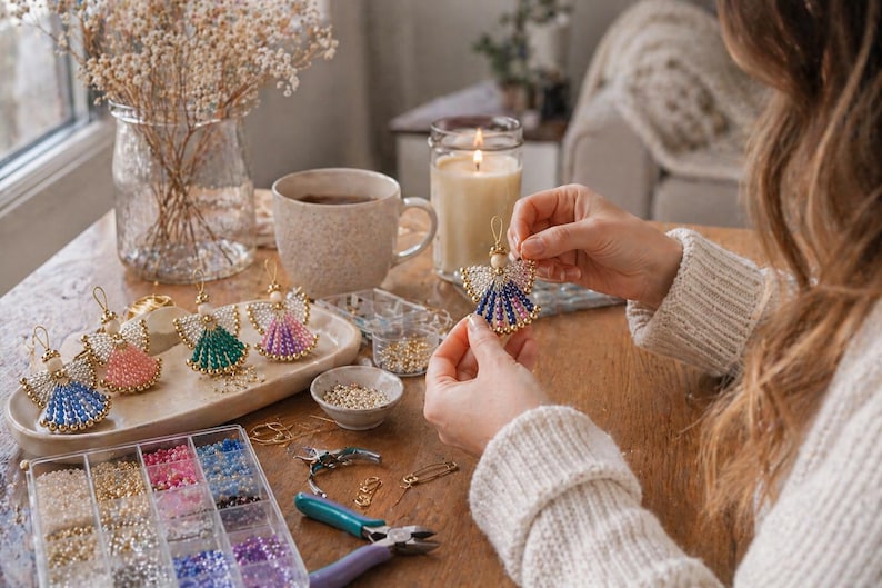 May include: A person is holding a beaded angel ornament with blue, pink, and gold accents. Other angel ornaments in various colors are displayed on a tray. Crafting supplies, including beads and tools, are visible on a wooden table.