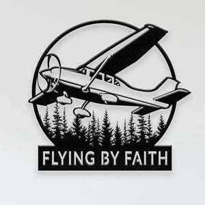 May include: Black metal wall art featuring an airplane design within a circular frame, above a forest silhouette. The text "FLYING BY FAITH" is displayed below the image. The artwork is mounted on a white wall.