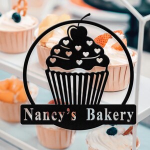 Könnte beinhalten: Schwarzes Metallschild in Form eines Cupcakes mit dem Text "Nancy's Bakery". Das Cupcake-Design zeigt eine Kirsche und herzförmige Ausschnitte. Im Hintergrund sind Cupcakes mit weißem Zuckerguss und Fruchtbelägen zu sehen.