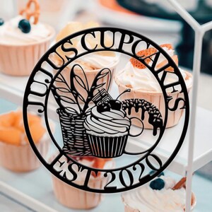 Könnte beinhalten: Schwarzes Metallschild mit den Worten "JULIUS CUPCAKES EST 2020" in einem kreisförmigen Design. Das Schild zeigt einen Cupcake, einen Korb und andere Illustrationen zum Thema Bäckerei. Cupcakes mit Zuckerguss und Toppings sind im Hintergrund zu sehen.