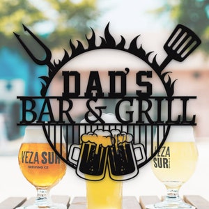 Könnte beinhalten: Ein schwarzes Metallschild mit dem Text "Dad's Bar & Grill" in einem Kreis mit Flammen und einem Grillspatel. Im Vordergrund stehen zwei Gläser Bier.
